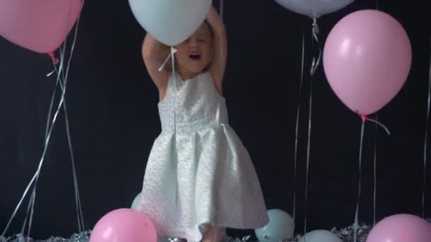 Un portrait d'une belle petite fille sourit et tient dans ses mains un ballon couleur en studio avec de nombreux ballons et une boutique de bonbons . — Video