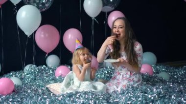 Sevimli küçük kız ve onun güzel genç anne yemek kek, doğum günü partisi, tatil partisi. Balonlar arka plan üzerinde