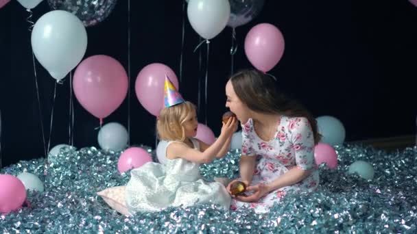 Menina bonito e sua bela jovem mãe comer cupcakes, festa de aniversário, festa de férias. No fundo dos balões — Vídeo de Stock