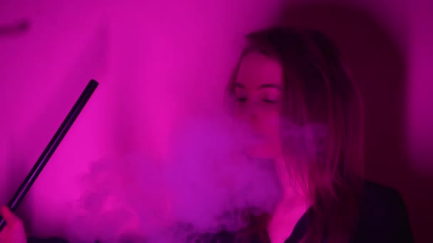 Mooie, jonge, hookah-rokende vrouw. Een aantrekkelijk meisje rookt tabak op smaak. Adem de rook uit in het neonlicht. — Stockvideo