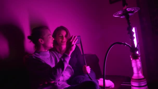 Ένας άνδρας και η κοπέλα του κάθονται σε ένα δωμάτιο hookah βλέποντας ένα βίντεο στο τηλέφωνό τους — Αρχείο Βίντεο