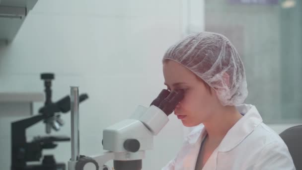 女性科学者は顕微鏡を使い始める。顕微鏡を探している微生物学者。微生物学研究室。研究室で顕微鏡を見ている女性科学者。顕微鏡. — ストック動画