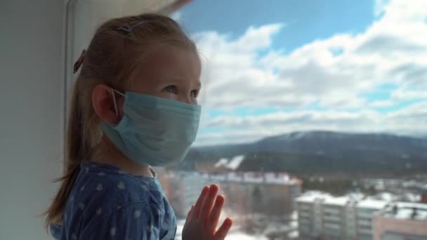 Niña con una máscara médica mirando por una ventana durante una epidemia mundial de virus. Detener la infección, la gripe y el SARS. Niño en aislamiento, cuarentena . — Vídeos de Stock