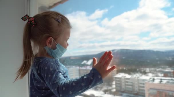 Uma menina com uma máscara médica a olhar pela janela durante uma epidemia de vírus mundial. Parem a infecção, a gripe e a SARS. Criança em isolamento, quarentena . — Vídeo de Stock