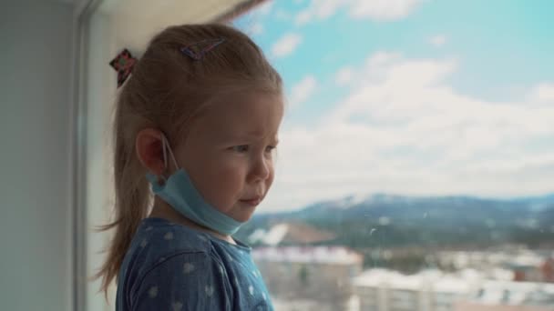 Kleines Mädchen in medizinischer Maske, das während einer weltweiten Virusepidemie aus dem Fenster schaut. Stoppt Infektionen, Grippe und SARS. Kind in Isolation, Quarantäne. — Stockvideo