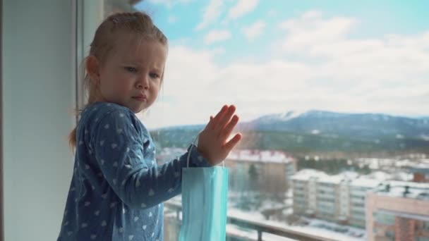 Niña con una máscara médica mirando por una ventana durante una epidemia mundial de virus. Detener la infección, la gripe y el SARS. Niño en aislamiento, cuarentena . — Vídeos de Stock