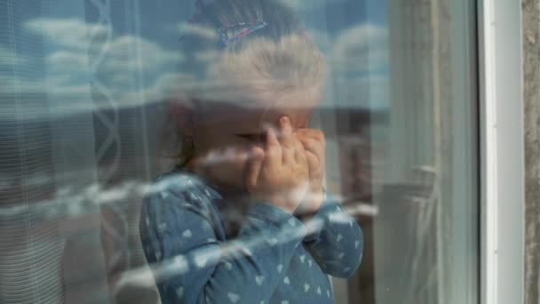 El niño está en casa aislado fuera de la ventana, llorando y quiere jugar afuera. Una niña golpea el cristal con su mano . — Vídeo de stock