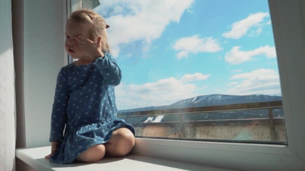 El niño está en casa aislado fuera de la ventana, llorando y quiere jugar afuera. Una niña golpea el cristal con su mano . — Vídeos de Stock