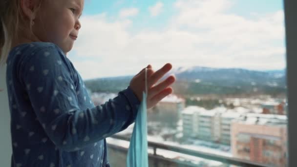 Κοριτσάκι με ιατρική μάσκα να κοιτάει έξω από το παράθυρο κατά τη διάρκεια μιας παγκόσμιας επιδημίας ιών. Σταματήστε τη μόλυνση, γρίπη και SARS. Παιδί σε απομόνωση, καραντίνα. — Αρχείο Βίντεο