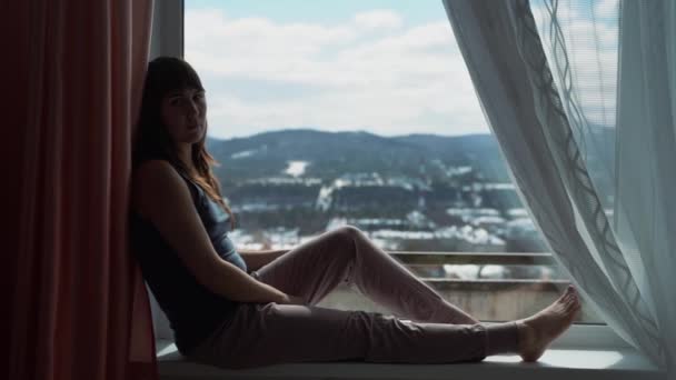 Mujer joven sentada en el alféizar de la ventana soñando despierto mirando al aire libre luz natural. Autoaislamiento, cuarentena — Vídeos de Stock