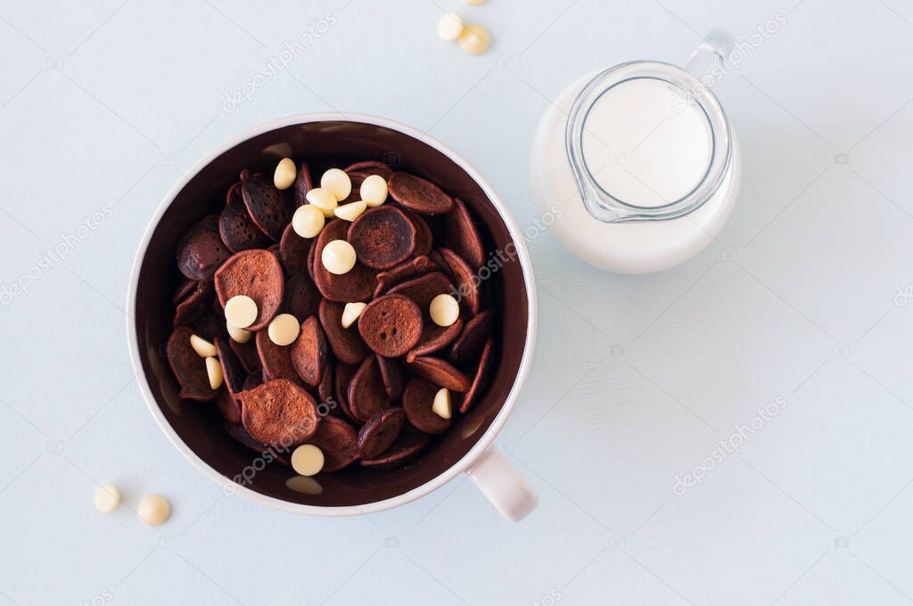 Chocolate tiny pancake cereal with white chocolate in a bowl and milk. Top view. Quarantine trendy food or new normal concept.