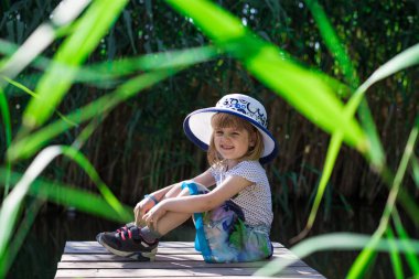 Şapkalı bir neşeli küçük kız nehir kıyısında oturur