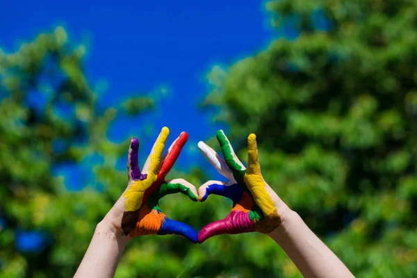 Las manos de los niños pintadas en colores brillantes hacen una forma de corazón en el fondo de la naturaleza de verano —  Fotos de Stock
