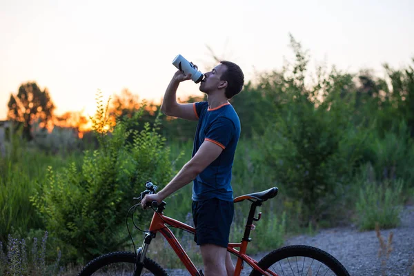 Молодий спортивний чоловік їде на велосипеді питною водою зі спортивної пляшки на заході сонця — стокове фото