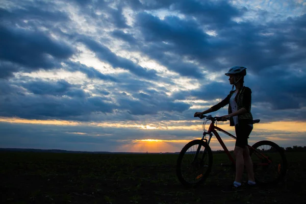 Silhouette einer jungen Frau mit Fahrrad im Feld bei Sonnenuntergang Himmel mit dramatischen Wolken — Stockfoto