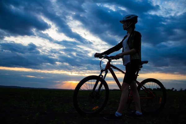 Силуэт молодой женщины с велосипедом в поле на закате неба с драматическими облаками — стоковое фото
