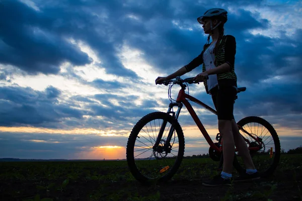 Silhouette einer jungen Frau mit Fahrrad im Feld bei Sonnenuntergang Himmel mit dramatischen Wolken — Stockfoto
