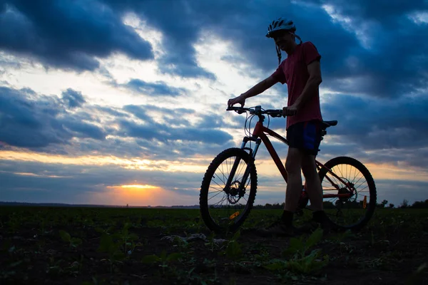 Silhouette eines jungen Mannes mit Fahrrad im Feld bei Sonnenuntergang Himmel mit dramatischen Wolken — Stockfoto