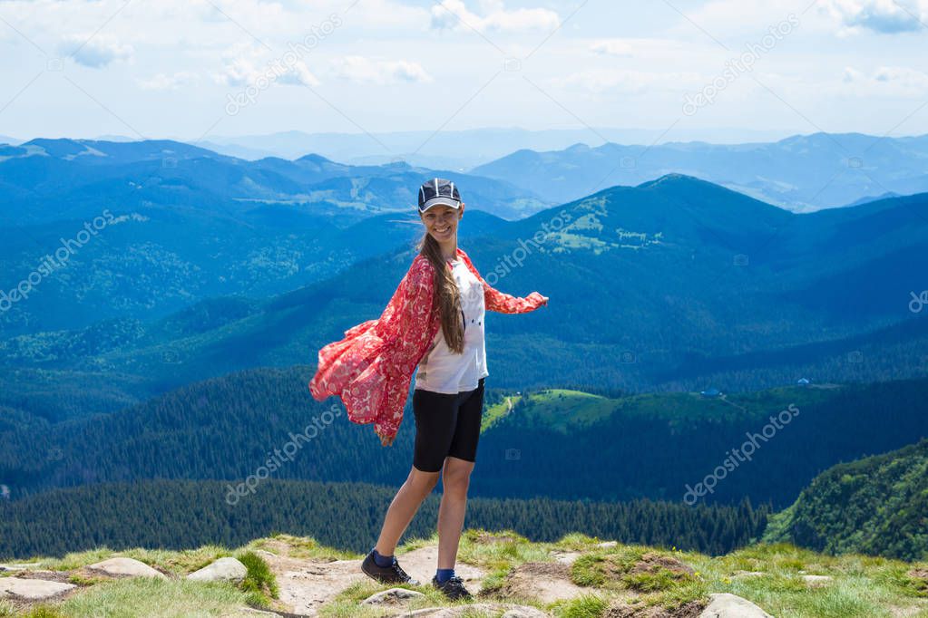 A happy young woman in red with hands up stands on top of a hill