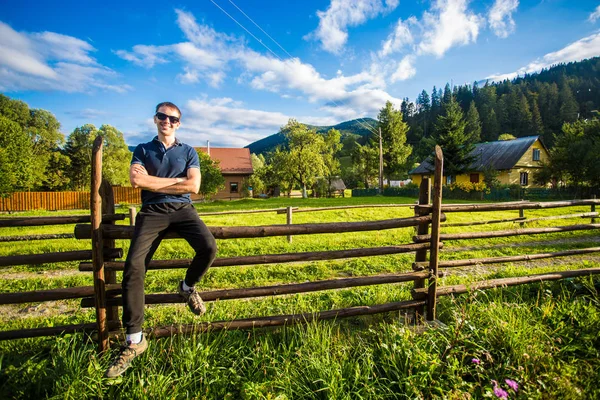 Молодой счастливый красивый мужчина в солнечных очках, отдыхающий на деревянном заборе в саду — стоковое фото