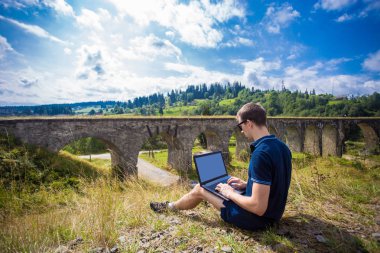 Dizüstü bilgisayar açık ve eski taş demiryolu Köprüsü yakınlarında çalışma ile oturan bir genç adam