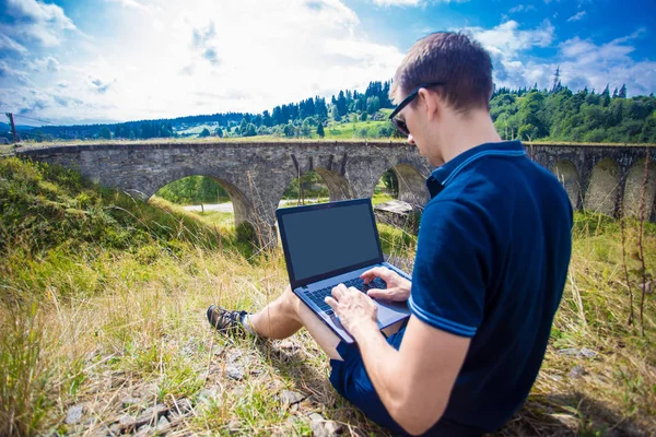 Молодой человек сидит с ноутбуком на открытом воздухе и работает возле старого каменного железнодорожного моста — стоковое фото