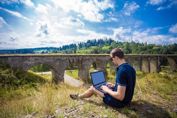 Dizüstü bilgisayar açık ve eski taş demiryolu Köprüsü yakınlarında çalışma ile oturan bir genç adam