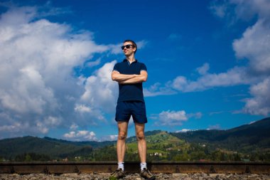 Güneş gözlüklü genç çekici adam mavi gökyüzüne karşı duruyor