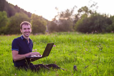 Dizüstü bilgisayar ile yeşil çimenlerin üzerinde oturan ve çalışan genç adam 