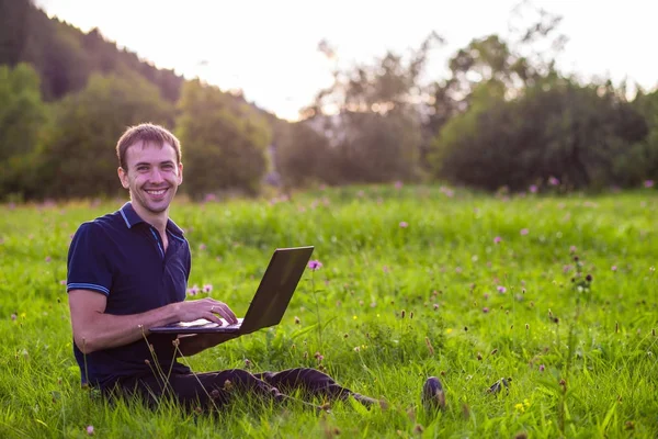 Dizüstü bilgisayar ile yeşil çimenlerin üzerinde oturan ve çalışan genç adam 