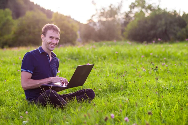 Dizüstü bilgisayar ile yeşil çimenlerin üzerinde oturan ve çalışan genç adam 