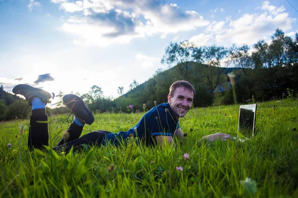 Молодой человек лежит на зеленой траве с ноутбуком и работает — стоковое фото