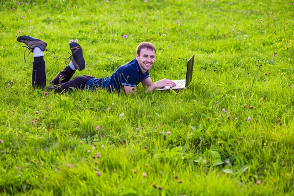 Молодой человек лежит на зеленой траве с ноутбуком и работает — стоковое фото