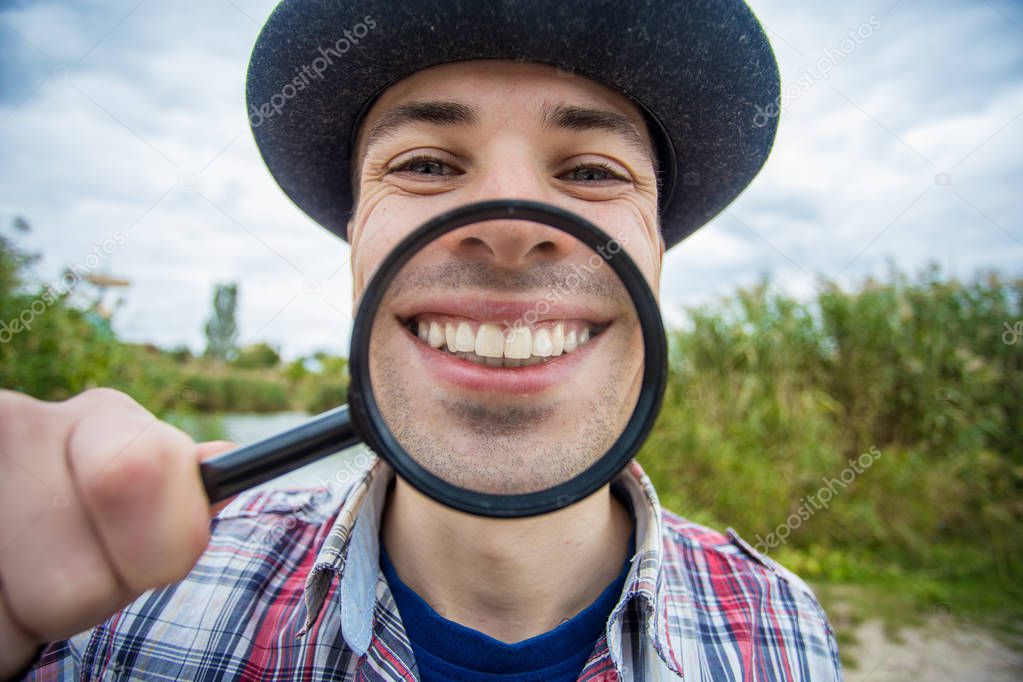 A cheerful man with a funny face in a hat holds a magnifying glass