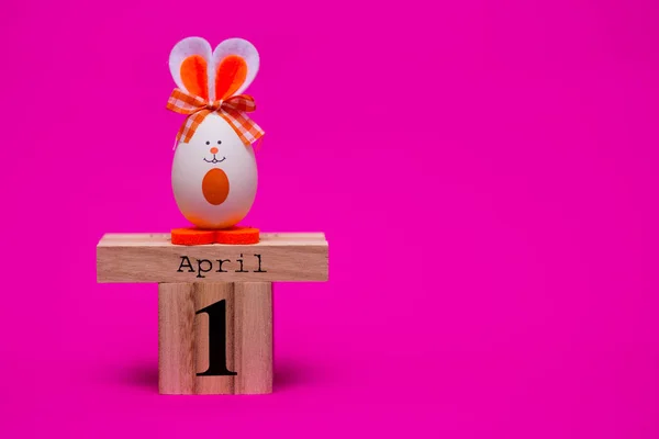 多彩多姿的滑稽的蛋以可爱的兔子的形式与木日历设置在4月1日被隔绝在粉红色背景 复活节快乐卡 愚人节 节日装饰 — 图库照片