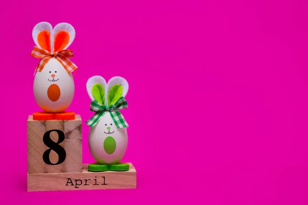 多彩多姿的滑稽的蛋以可爱的兔子的形式与木日历设置在4月8日被隔绝在粉红色背景 复活节快乐卡 节日装饰 — 图库照片