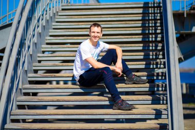 Genç adam merdivenlerde güneşli bir günde oturuyor. Sokak stili