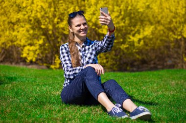 Güzel kız Park'ta rahatlayın ve selfie tree yakınındaki yeşil çimenlerin üzerinde sarı çiçek al. Bahar zamanı. İletişim kavramı