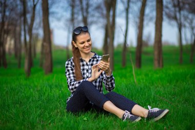 Mutlu kız çimlerin üzerine parkta oturan sms tipik. Kadın telefonda sohbet. Smartphone üzerinden ağlardan haberleri okuyun. Genç kadın telefona mesaj gönderme 
