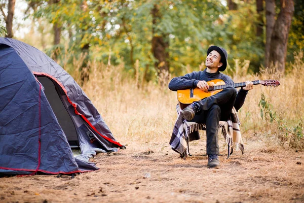 Joven sonriente con sombrero negro sentado cerca de una tienda turística y tocando la guitarra en el bosque — Foto de Stock