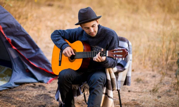 Joven sonriente con sombrero negro sentado cerca de una tienda turística y tocando la guitarra en el bosque — Foto de Stock