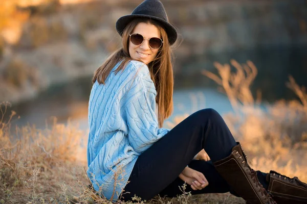 Retrato de menina bonita em óculos de sol e chapéu preto e suéter azul sentado perto do lago — Fotografia de Stock