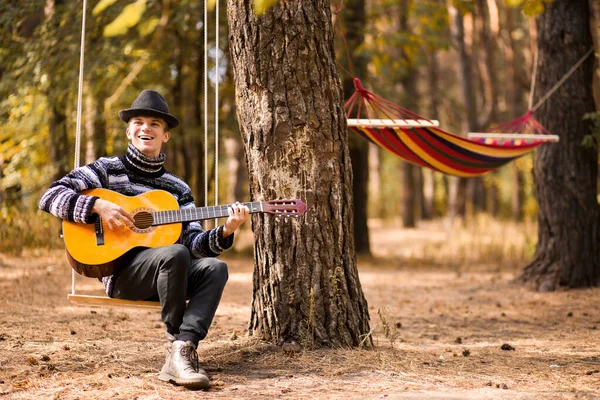 Joven hombre atractivo guapo en suéter tocando la guitarra en el bosque sentado en swing. Hombre casual, estilo de vida . — Foto de Stock