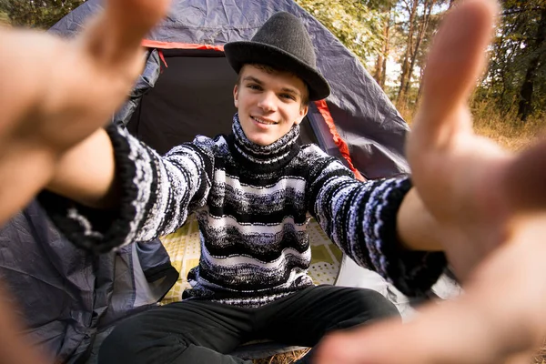 Joven sonriendo y tomando selfie en el campamento forestal desde el teléfono inteligente. Hombre viajero con sombrero negro tomar autorretrato vie teléfono móvil — Foto de Stock