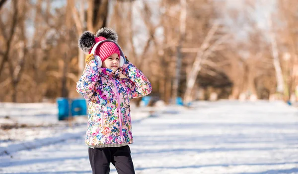Smiling little girl in pink hat with bubo walk in snow park and listen to music in headphones — Stock Photo, Image