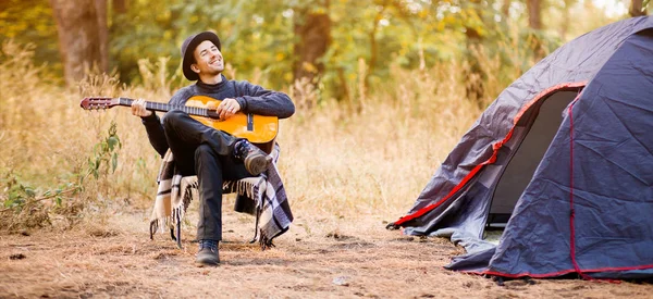 Siyah şapkalı gülümseyen bir gencin turistik çadırın yanında oturup ormanda gitar çalması.