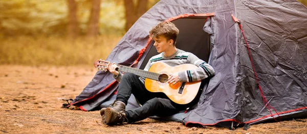 Banner vista de un joven sonriente sentado cerca de una tienda turística y tocando la guitarra en el bosque — Foto de Stock