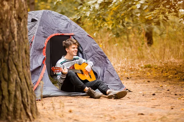 Sonriente joven sentado cerca de la tienda turística y tocando la guitarra en el bosque — Foto de Stock