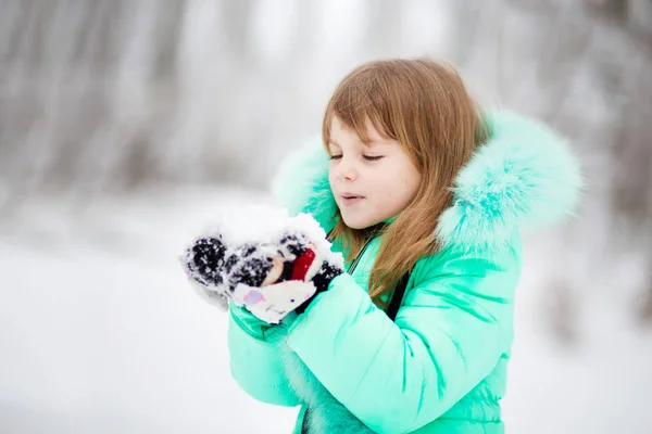 Маленькая девочка дует снегом с варежками в красивом парке во время снегопада — стоковое фото