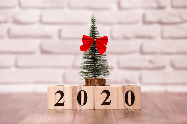 New year. Text 2020 on wooden calendar blocks with little christmas tree and red bow on white brick wall background.